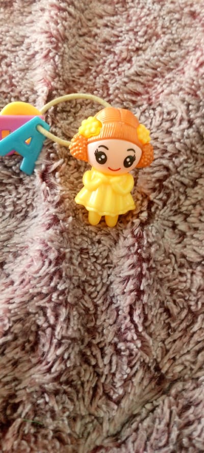 An overhead shot shows a small plastic doll standing on a fuzzy, brown and pinkish-red carpet. The doll has orange hair styled in two pigtails with yellow flower accents. It wears a yellow dress and has large black eyes and a red mouth. To the left of the doll, a blue plastic letter 'A' is attached to a yellow string. The doll and the letter 'A' appear to be part of a larger toy or decoration, possibly a garland. The background is entirely composed of the textured carpet.