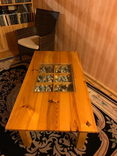 In the city of Mepiskalaki, Georgia, a scene unfolds within what appears to be a living room or study. The focal point is a wooden coffee table with a glass top, beneath which is a grid of compartments filled with shells and stones. The table has a warm, varnished finish and rests on a patterned rug with black and beige motifs. To the left, a woven wicker chair with a light-colored cushion sits next to a wooden bookshelf filled with books. The wall behind the chair is a warm, peachy-orange color, exhibiting a textured, possibly stuccoed finish with subtle vertical striations. The lighting in the room is warm, casting soft shadows. The overall atmosphere is cozy and suggests a personal, curated space.