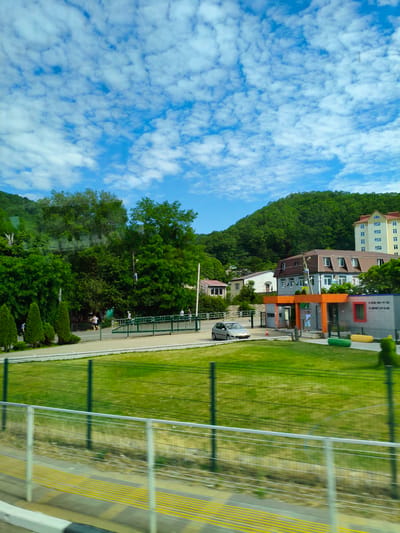 The image captures a scene in Agoy, Russia, with a focus on a verdant hillside dotted with buildings and lush trees under a bright blue sky filled with white, scattered clouds. In the foreground, a green metal fence runs horizontally, separating a paved area with yellow tactile paving from a neatly mown green lawn. Beyond the lawn, another green fence encloses a wider expanse of grass.
Further back, a road curves gently, with a light-colored car parked to the left. Several people are visible walking and standing near a green railing and a building with an orange-trimmed entrance. The building has modern architectural elements, with windows and what appears to be a reception area. Behind this building, a multi-story residential building with a light-colored facade and a brown roof stands prominently. Further up the hillside, more trees and smaller buildings are visible, blending into the natural landscape. The overall impression is of a well-maintained area within a hilly, natural environment.