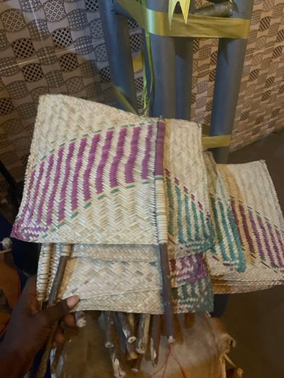 The image displays a close-up view of several intricately woven items, likely traditional fans or mats, made from natural fibers such as straw or dried palm leaves. These items are predominantly light beige or natural brown in color, and many feature decorative woven patterns in shades of purple, teal, and a darker beige. The weaving technique visible includes diagonal and herringbone patterns, showcasing skilled craftsmanship.
A collection of these woven items is gathered together, with one dark-skinned hand visible in the lower right, holding several of the wooden stick handles that are attached to the woven pieces. These handles appear to be made from rough-hewn wooden branches. To the right of the hand, more similar wooden sticks are stacked or arranged.
In the background, to the left, there is a patterned surface, possibly a floor or wall covered with a mat or textile. The pattern consists of repeating geometric shapes, resembling interconnected circles or ovals. Above this patterned surface, several grey cylindrical objects are partially visible, possibly poles or pipes, with some gold and green strips (perhaps ribbons or leaves) draped over them. In the upper right background, a blurred, light-brown, textured object, possibly a traditional container or drum made from animal hide, can be seen.
The scene suggests an environment where traditional crafts are made, displayed, or used, emphasizing natural materials and manual artistry. No specific text is visible in the image.