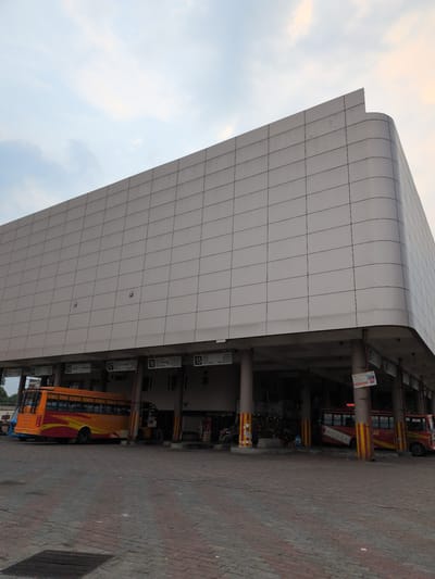 The image shows the Una, India bus stand. The bus stand features a modern, multi-story building with a large, tiled facade. In the foreground, a bright orange and yellow bus is parked. The bus has the words "HIMACHAL ROAD TRANSPORT CORPORATION" written on its side. A red and white bus is parked behind it. The ground is paved with brick-like tiles, and a metal grate is visible in the lower left corner. The sky above is overcast with hints of orange and pink from the setting sun. The overall scene depicts a functional transportation hub with elements of modern architecture.
