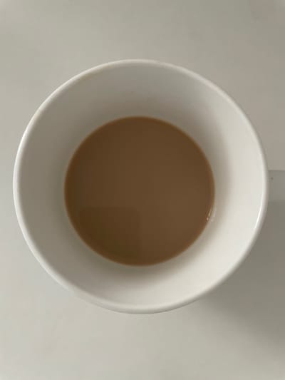 The image presents a top-down view of a white ceramic mug, centrally framed against a plain, light grey or off-white surface. The mug contains a light brown liquid, consistent in color with coffee or tea mixed with milk, filling approximately one-quarter of its volume. The surface of the liquid is smooth and still. The interior and exterior of the mug are uniformly white, though subtle, light brown discoloration or staining is visible along portions of the upper rim, particularly on the top-left edge. The surrounding surface is unadorned and lacks any identifying features. No individuals, additional objects, actions, or contextual elements are depicted. There are no visual cues or landmarks within the image that allow for the determination of a specific geographical location, including the city of Емельяново, Russia.