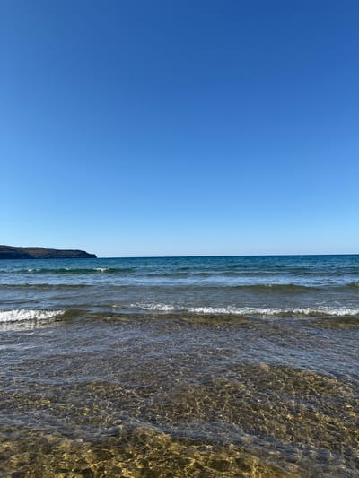 This is a landscape photograph of a clear blue sky above a calm sea. Gentle waves break on a shallow, rocky shore. In the distance, a dark, tree-covered headland juts out into the sea. The image was taken in Káto Stalós, Greece. There are no people or man-made objects visible in the scene.