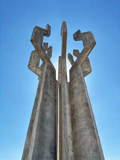 A low-angle shot shows a weathered, concrete abstract sculpture against a clear, light blue sky. The sculpture features geometric shapes and intersecting beams, revealing sharp angles and rough surfaces. The sunlight peeks through the gaps in the structure, creating a glowing effect. The sculpture has a horizontal orientation in the frame. The context indicates that it is located in Barutana, Montenegro.