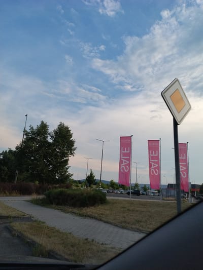 The image depicts an outdoor scene with a cloudy blue sky above. In the foreground, a paved walkway leads into a grassy area. To the left, there are trees and bushes. Further in the background, there is a parking lot with several cars. Three tall pink flags with white text are prominently displayed, and a diamond-shaped road sign is visible on the right side of the frame. The scene appears to be a commercial area, possibly a shopping center or retail park. The overall atmosphere is calm and suggests daytime.