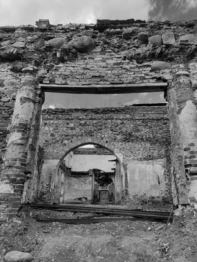 The image presents a black and white view of a ruined structure in Жалудоцкі сельскі Савет, Belarus. The building appears to be constructed from a combination of brick and stone. There are visible openings, including a window and an archway, which reveal further interior deterioration. The environment is marked by rubble and debris.