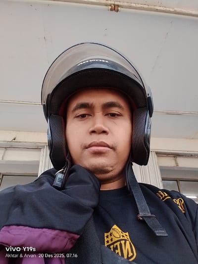 A man wearing a black helmet with a clear visor looks directly at the camera. The helmet's brand, "HONDA," is visible. He is wearing a black long-sleeved shirt with a gold emblem and accents. Part of a dark jacket with a purple lining is also visible on the left side. The background consists of an off-white wall, a window, and a door frame. The lower left corner displays "vivo Y19s Akhtar & Arvan 20 Des 2025, 07.16."