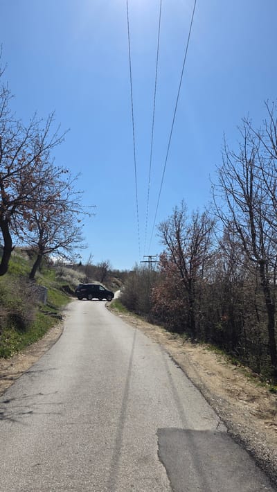 The image shows a narrow paved road curving through a rural area near Elenovo, Bulgaria. A dark-colored hatchback vehicle is parked on the road, facing away from the viewer. The road is bordered by dry vegetation and trees with bare branches. Overhead power lines stretch across the sky. The sky is clear and blue. There are no people visible in the image.