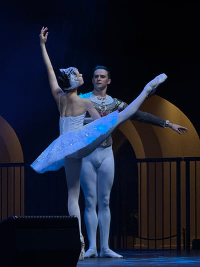 A male ballet dancer holds a female dancer horizontally, both wearing white tights and costumes. The woman is in a white tutu with a feathered headpiece, arms extended. The man, adorned in a decorative tunic, supports her outstretched leg. The backdrop features a dark stage with gold and black geometric designs. The scene captures a moment of artistic performance and collaboration.