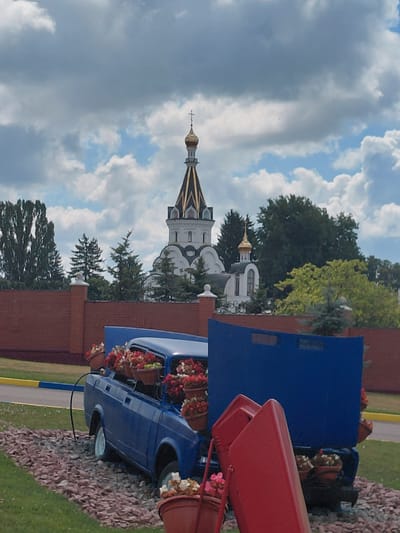 The image depicts an outdoor scene featuring a blue car repurposed as a planter. The car is filled with numerous potted flowers, primarily red and white, overflowing from its interior, engine compartment, and trunk. The car is situated on a bed of reddish-brown gravel, with green grass bordering the foreground. In the background, a brick wall runs horizontally, with a decorative post visible on the left. Beyond the wall, a white building with golden domes and a cross on top, characteristic of an Orthodox church, stands prominently against a partly cloudy sky. Lush green trees surround the church and the wall. The scene appears to be a decorative installation in a public space, possibly near the church. The location is identified as Kovalivka, Ukraine. There are no people visible in the image.