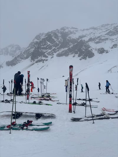 The scene depicts a snow-covered alpine environment in Klösterle, Austria, under overcast skies with visibility diminishing towards the distant, rugged mountain peaks. The foreground and mid-ground consist of an expansive snowfield serving as an active ski area.
Approximately 10 to 15 individuals are present, all dressed in full winter ski attire, including jackets, pants, helmets, and goggles. Numerous pairs of skis and ski poles are scattered throughout the snow; many skis are planted vertically, while others lie horizontally. One snowboard is clearly visible, resting flat on the snow in the lower-left foreground.
In the left-center, two individuals are standing near a cluster of upright skis; one person in a dark jacket and blue helmet appears to be interacting with or adjusting equipment. A person in an orange jacket is positioned slightly behind them. Other individuals are distributed across the snowfield; some are standing stationary, while others are in active motion on their skis, such as a person in a blue jacket on the right side of the frame, actively skiing downhill with poles. The overall environment is a high-altitude, snow-covered mountain slope.