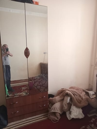 The image captures an indoor scene, likely a bedroom, with a large mirrored wardrobe dominating the foreground. The wardrobe has multiple doors, two of which are clearly visible and reflect the surroundings. In the mirror, a person is seen lying down and holding a smartphone to take a picture of themselves, a selfie. The person is wearing a light-colored t-shirt and dark pants.
To the right of the wardrobe, a wooden dresser with several drawers is visible, each with a silver handle. The top of the dresser is cluttered with a large, fluffy teddy bear and other soft items, possibly blankets or clothes, in shades of brown and white. The floor is covered with a rug featuring a red and beige pattern. The walls appear to be a light cream color.
Above the wardrobe, a white ceiling with some piping or conduit is visible. The overall lighting suggests it is daytime, with natural light likely entering from an unseen window. There is no visible text in the image, except for the partial word "OR" on an orange object partially visible on the far left edge of the image. The scene appears casual and personal, suggesting a moment of quiet activity within a home. The presence of the teddy bear adds a touch of coziness or perhaps indicates the presence of a child or someone who enjoys stuffed animals. The setting is consistent with a bedroom or a dressing area.