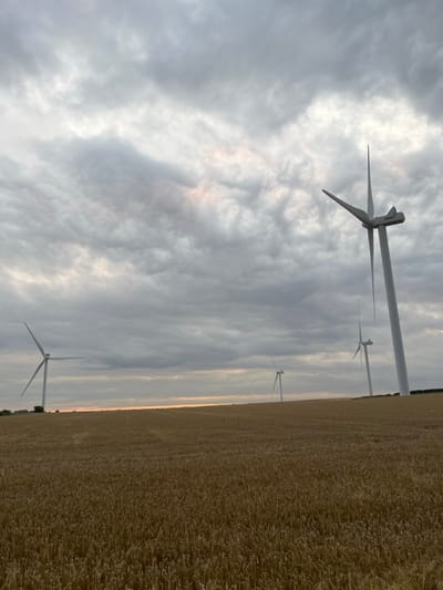 The image depicts a rural landscape near Barmston, United Kingdom, under an overcast sky. A vast field of mature, golden-brown grain, likely wheat or barley, occupies the lower half of the frame and extends to the distant horizon. Four prominent white wind turbines are visible; one large turbine stands partially in the foreground on the right, with its blades static. Three additional turbines are positioned further into the distance across the mid-ground, appearing progressively smaller. All visible turbine blades are stationary. The sky is filled with dense, grey clouds, showing a faint band of orange-pink light along the low horizon line. No individuals, animals, or vehicles are present within the scene.