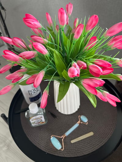This is an overhead shot of a dark brown woven placemat on a black round table. The placemat holds several objects, suggesting a skincare or beauty routine setup. In the foreground and to the left, a large bouquet of artificial pink tulips with green stems and leaves dominates the frame. To the right of the flowers, a white fluted object, possibly a vase or decorative container, is visible. Further to the right, a light blue jade facial roller with rose gold accents lies on the placemat, alongside a thin, light beige implement that could be a cosmetic tool. Below the facial roller, a small clear glass bottle with a white cap contains a clear liquid labeled "Coconut Oil" and features an image of a coconut. A white bottle with blue lettering, partially obscured by flowers, reads "Podi Logic." The setting appears to be indoors, with natural light illuminating the scene. The overall mood is calm and organized, hinting at a moment of self-care. The location is identified as Rubtsovo, Russia, though no specific geographical cues are present in the image itself.