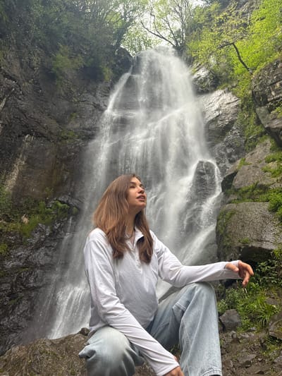 A woman is photographed in front of the Makhuntseti Waterfall, located in Kveda Makhuntseti, Georgia. The woman has brown hair and is wearing a white long-sleeved shirt and light blue jeans. She is positioned on rocks, looking downward towards the waterfall. The waterfall itself is cascading down a rocky cliff face, with some greenery visible on the rocks surrounding the waterfall. The scene is set in a natural environment.