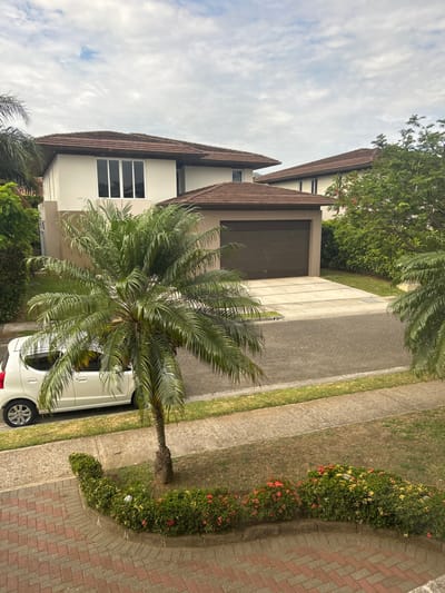 The image shows a residential street in Río Oro, Costa Rica (latitude: 9.945, longitude: -84.201). In the foreground, a white car is parked on the right side of the street. A large palm tree stands between the car and the house. The street is paved with grey concrete blocks and bordered by lush green grass. A small, manicured flowerbed with red blossoms is in the lower right corner of the image. The houses in the background are all modern, single-story buildings with tiled roofs. They are painted white and have large windows. There is a garage in front of one of the houses, with its door closed. The image was taken from a slightly elevated viewpoint, looking down onto the street. The weather is clear and sunny, with some clouds in the sky.