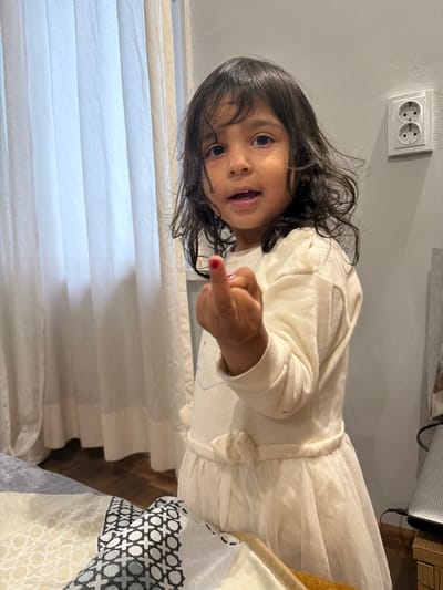 A young child, possibly female, with dark hair and dark eyes, is standing in what appears to be a domestic indoor setting. The child is wearing a cream-colored dress with a ruffled skirt and has red nail polish on their right index finger. The child's right hand is raised, with the index finger extended, in a gesture that could be interpreted as defiant or playful.
In the background, a white curtain is visible on the left, suggesting a window. On the right side of the image, a light gray wall is present, with a double electrical outlet mounted on it. The lighting in the scene appears to be natural, possibly from the window. The lower part of the image shows a patterned duvet or bedspread.
The overall environment suggests a bedroom or living area. The image does not contain any elements that would specifically place it in the city of Dalgopol, Bulgaria.