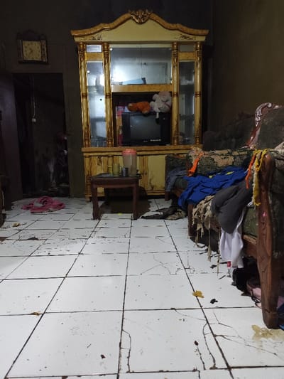 The image shows the interior of a home located in Klapanunggal, Indonesia, at the coordinates provided. The home is sparsely furnished, with a large wooden cabinet with glass doors in the center of the room. The cabinet is filled with various items, including a small television and some stuffed animals. A small table with four chairs sits in front of the cabinet, and a large couch sits in the right side of the image. Several articles of clothing are draped over the couch, and a pair of shoes are on the floor near the couch. The floor is tiled and dirty, and there are several cracks in the tiles. The room is dark and cluttered.