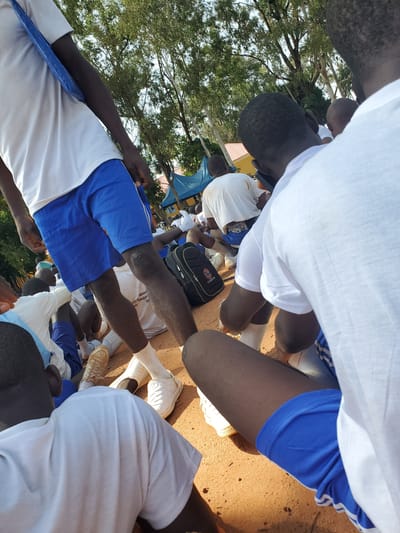 In Barakin Rafin Gora, Nigeria, a group of young people, likely students, are gathered outdoors on a dirt surface, possibly a schoolyard. The scene is partially obscured by the perspective, focusing on the lower bodies and the ground. The individuals are wearing white t-shirts and blue shorts, some with white stripes. Several are sitting or kneeling, with some visible wearing white socks and sneakers. A black backpack with a white logo is on the ground near some of the individuals. The background shows trees with lush green foliage and a glimpse of a red-roofed building and a blue tent. The lighting suggests it is daytime, with sunlight casting shadows on the ground.