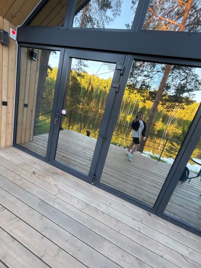 A person, appearing female, dressed in a white t-shirt, black jacket, black cycling shorts, and light blue athletic shoes, is reflected in large glass doors and windows of a modern building in Mishkino, Russia. The individual is positioned on a weathered wooden deck, holding a smartphone to capture the reflection.

The building features dark gray frames for the floor-to-ceiling glass panels, with a light-toned wooden facade on the left. A prominent white sticker with black Cyrillic text, "НАЦИОНАЛЬНЫЕ ПРОЕКТЫ РОССИИ" (NATIONAL PROJECTS OF RUSSIA), is affixed to an upper glass panel. A small pink circular sticker is on the handle of the central door, which includes a keyhole. Exterior elements visible on the wooden facade include a square black electrical box, a red light sensor, and white conduit. Further inside the reflection on the left, a black speaker or similar device is mounted on the wooden wall.

The reflection reveals a black metal railing separating the deck from a calm body of water, likely a lake or river, which stretches towards a dense forest on the opposite bank. The forest exhibits a mix of green foliage and lighter-barked trees, illuminated by what appears to be late afternoon or early morning sunlight. The sky, visible through the upper glass, is clear blue. A dark backpack and part of a table are visible on the deck in the reflection on the right side. The wooden deck itself shows signs of wear and some minor discoloration.