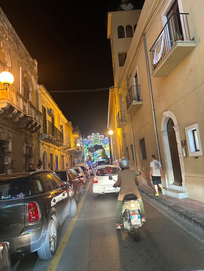 The image depicts a night scene on a narrow, illuminated street in Giardini-Naxos, Italy. Multi-story buildings with architectural details and balconies line both sides of the street. Street lighting emanates from spherical yellow fixtures. A clock tower with a visible clock face stands in the upper background on the right.
A line of vehicles, including a dark Mini Cooper in the left foreground and a white hatchback, occupies the central street lane. In the foreground center, a person wearing a gray helmet, brown coat, and green pants operates a beige scooter with Italian license plate SP 41852, positioned facing away from the camera. The scooter operator wears red-accented shoes.
On the right sidewalk, a male pedestrian wearing a light t-shirt with a graphic, dark shorts, and yellow footwear walks away from the camera's perspective. In the distant background, a large, multi-colored illuminated archway spans the street, visible between the buildings. Multiple individuals are discernible on the left sidewalk, some near parked cars. One balcony on the right building displays a hanging banner.