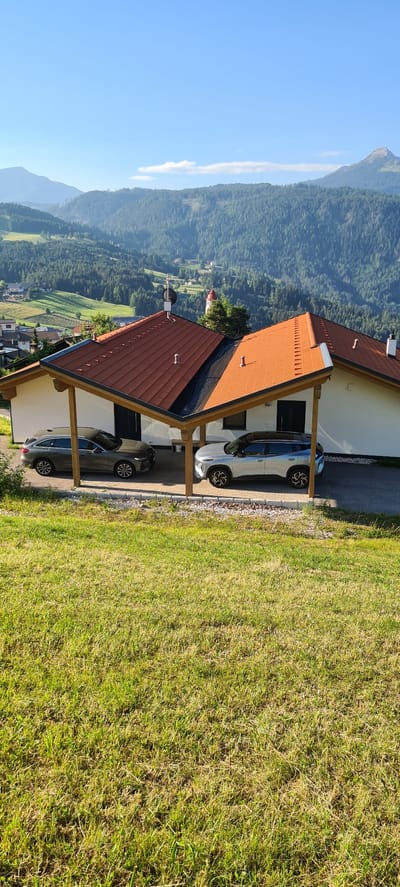 This image showcases a scenic mountain landscape in Deutschnofen - Nova Ponente, Italy, under a clear blue sky with a few wispy clouds. The foreground features a modern house with a red-tiled roof and a covered carport. Two cars are parked under the carport: a silver electric car and a dark gray sedan. A well-maintained lawn with patches of dry grass borders the house.

In the background, lush green forests cover the rolling hills, leading up to majestic mountains. The scene appears to be set in the late afternoon, indicated by the warm sunlight casting shadows. The overall atmosphere is peaceful and serene, highlighting the natural beauty of the alpine region. No people or text are visible in the image.