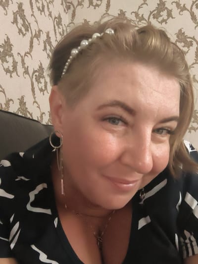 This is a close-up, self-portrait style photograph of a woman with blonde hair and a pearl headband. She is wearing a black top with a white abstract pattern and a silver dangle earring. Her eyes are blue-green and she has light-colored skin. The background appears to be wallpaper with a vintage floral pattern. The image is taken indoors and the lighting is warm and soft. The location is not specified in the image or the provided context.