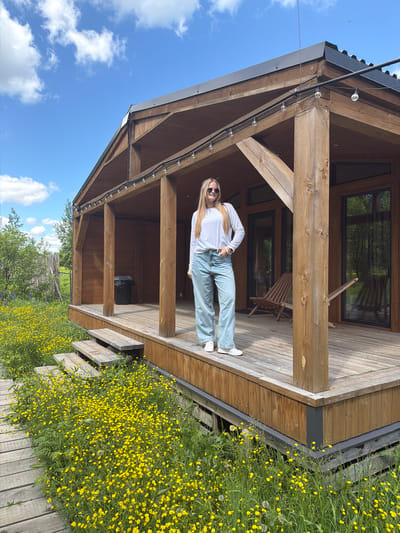 This outdoor photograph, taken in Vozhoiskii, Russia, captures a bright, sunny day featuring a young woman posing on the wooden porch of a cabin or house. The scene is dominated by natural elements and rustic architecture.

The central subject is a young woman with long, blonde hair, wearing sunglasses, a white long-sleeved shirt, light blue wide-leg jeans, and white sneakers. She stands casually on the porch, one hand on her hip, with a faint smile directed towards the viewer, appearing relaxed and happy.

The porch is constructed from warm-toned wooden planks and features prominent wooden support beams. Decorative string lights with clear bulbs are strung along the underside of the porch roof. Through a large window on the back wall of the porch, a wooden lounge chair is visible, along with reflections of surrounding trees. A black trash can or similar container is also present on the porch floor.

The area immediately surrounding the cabin is a vibrant meadow of lush green grass densely covered with countless small yellow flowers, likely buttercups, and some dandelions. A rustic wooden walkway or steps extends through this floral landscape. In the distance, trees and what appears to be a fence line the horizon. The sky is a clear, bright blue with scattered white clouds, indicating fair weather and likely mid-day sunshine. There is no visible text in the image.