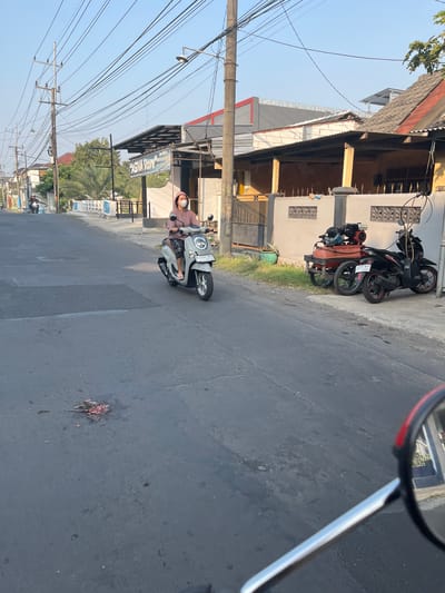 The image depicts a street scene in Sugih Waras, Indonesia, during daylight. In the foreground, a person wearing a pink hooded jacket and a white face mask rides a light-colored scooter. The scooter has a license plate reading "W 3764 NGO". The rider is wearing checkered pants and flip-flops. On the asphalt road in the lower left quadrant of the image, there is evidence of a deceased animal, with some scattered blood and body parts.
To the right of the road, a black motorcycle is parked next to a red air compressor on a cart. Behind these items is a light-colored wall with decorative patterns. Further back, a building with a dark-colored roof and supporting pillars is visible. A sign with the text "AGIVA Store" is mounted on the building.
The environment includes a paved road, sidewalks, buildings, utility poles with numerous wires, and some vegetation, including palm trees. The sky is clear and blue, suggesting a sunny day. The overall impression is of a quiet residential or commercial street.