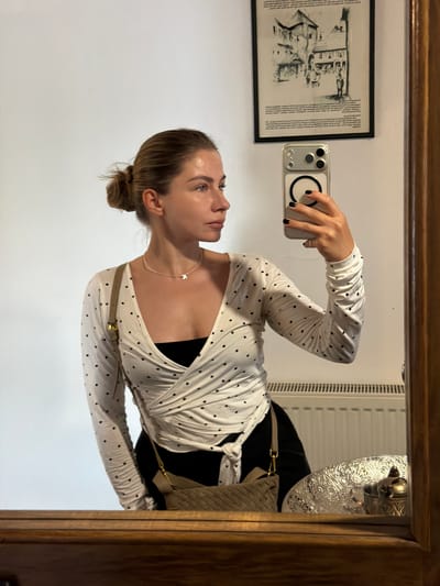 The image, taken in Ćusine, Bosnia and Herzegovina, shows a woman in a mirror's reflection. She is wearing a white, long-sleeved top with black polka dots, a black bra underneath, and dark trousers. A beige crossbody bag with gold hardware is slung over her shoulder. She is holding a smartphone in her right hand, which is angled to capture the reflection. A delicate necklace with a small charm is visible around her neck. Her hair is pulled back in a bun.
In the background, visible in the reflection, is a framed print on the wall, depicting a black and white drawing of buildings and people. To the right, a decorative metallic object is mounted on the wall, and a radiator or ventilation system is visible. The overall setting appears to be an indoor space, possibly a home, with a wooden frame surrounding the mirror. The lighting suggests it could be daytime, given the brightness of the reflection. The woman's expression is neutral, as she focuses on taking the photograph.