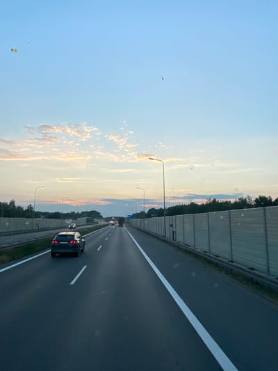 The image depicts a highway scene in Podłęże, Poland, during dusk. The sky is a clear blue with scattered clouds tinged with orange and pink from the setting sun. A road stretches into the distance, divided into multiple lanes by white lines. Cars are visible on the highway, with one dark SUV in the foreground and a truck further ahead. On either side of the road, tall, beige sound barriers are installed, with trees and foliage visible beyond them. Streetlights are spaced along the highway. The overall impression is one of a typical evening commute on a Polish highway.