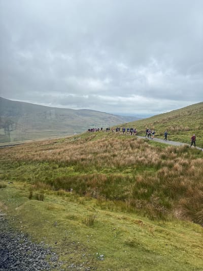 The image depicts a group of people hiking on a grassy hillside. The path they are following is narrow and well-worn. The hillside is steep, with varying shades of green and brown grasses. In the distance, there are more rolling hills and a cloudy sky. The photograph appears to be taken in the mountains surrounding Llanberis, United Kingdom.