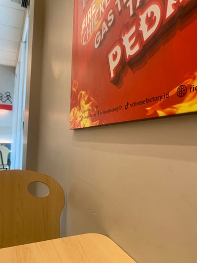 The image displays an interior view of a Richeese Factory restaurant, identified by its branding on a prominent wall poster, located in Calukan RT 1 RW 5 Keboansikep, Indonesia. The immediate foreground features the corner of a light-toned wooden or laminate table. Behind the table and to the left, the top portion of a matching wooden chair with an oval cutout in its backrest is visible. A pale beige wall occupies the majority of the right and central frame. Mounted on this wall is a large, rectangular red poster with orange and yellow flame graphics at its bottom edge. White text on the poster reads "FIRE CHICKEN," "GAS," and a large stylized "PEDAS." Below this, social media handles "@richeese_factory" and "richeesefactory.id," along with a TikTok icon labeled "richeesefactoryID," are present. Small text also states "Contoh hanya ilustrasi." To the far left, a white-trimmed doorway or opening reveals a brightly lit area beyond, where a red surface with black stick-figure graphics is partially discernible. No individuals, direct actions, or interactions are present in the static scene.
