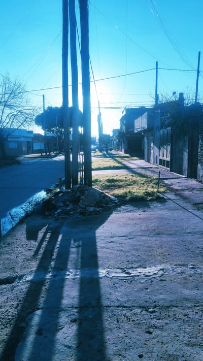 A low-angle, close-up shot captures a sidewalk in Remedios de Escalada, Argentina, under a bright blue sky. The foreground is dominated by the textured concrete of the sidewalk, showing cracks and patches of wear. To the left, a pile of rubble, consisting of broken concrete and debris, sits near the edge of the sidewalk. Three utility poles, silhouetted against the bright sky, stand prominently in the middle ground. Their shadows stretch long across the sidewalk and a patch of grass and weeds to the right. In the background, a residential street is visible, lined with houses and buildings. The sun is low, creating a strong backlight effect and casting long shadows. The overall impression is one of a quiet, early morning or late afternoon in a residential neighborhood.
