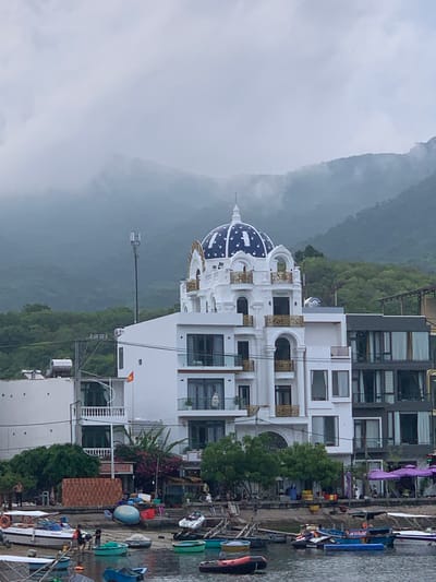 The image shows a waterfront scene in Vĩnh Hải, Vietnam, with a focus on a white building featuring a distinctive blue and white domed roof with gold accents. The building has multiple stories with numerous windows and balconies. To the right of the building, several boats are docked in the water, some appearing to be small fishing or tour boats. The water is calm, reflecting the overcast sky.
In the background, lush green mountains rise, partially obscured by mist or clouds, suggesting a humid or rainy climate. The weather appears to be overcast and possibly damp, as indicated by the muted lighting and the slightly hazy atmosphere.
The scene includes some people visible on the dock and possibly on the boats, but they are not the primary focus. There are also various objects like containers, umbrellas, and signage on the dock. One sign is partially visible with Vietnamese text that appears to read "VỊNH HẢI MINH BAY". The overall impression is a somewhat serene, albeit cloudy, day in a coastal or riverside town. The architectural style of the main building, with its dome and ornate details, stands out against the natural backdrop of the mountains.