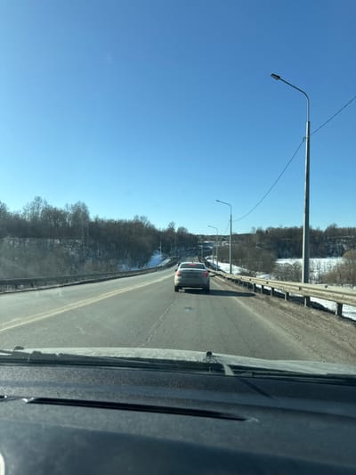 The image depicts a forward-facing view from inside a moving vehicle on a multi-lane asphalt road, consistent with a location in Ярославка, Russia. The lower portion of the vehicle's dark dashboard and windshield wipers are visible. Ahead, a light-colored sedan is traveling in the same direction on the right-hand lane. The road is flanked by metal guardrails on both sides. To the left, an embankment rises, covered with bare deciduous trees and patches of snow on the ground. To the right, the terrain slopes downwards, also featuring bare trees and snow. Multiple streetlights with black fixtures on tall silver poles line the right side of the road, connected by overhead power lines. The sky is clear and bright blue. The bare trees and snow indicate a winter or early spring season. No individuals are visible.