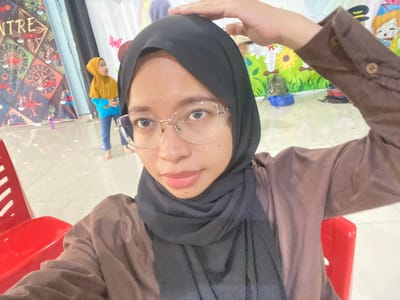 A woman in a black hijab and brown top is taking a selfie. She is wearing glasses and has her left hand on her head. In the background, a young child in a yellow hijab and blue shirt is standing. The scene appears to be indoors, possibly at a community center or event space in Tanjung Gading, Indonesia, as indicated by a banner with the word "NTRE" visible on the left, and a brightly colored mural depicting cartoon characters and sunflowers on the right. Red chairs are present in the foreground.