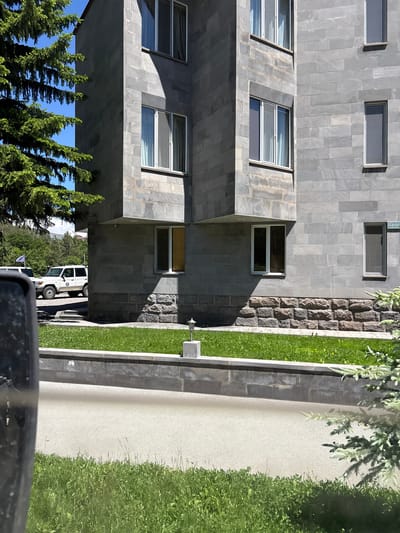 The image shows a modern building with grey stone facade and several windows. In front of the building, there is a well-maintained green lawn with a short stone wall bordering a paved area. A white SUV is parked on the paved area. A large evergreen tree is partially visible on the left side of the image. The sky is clear and blue. On the building facade, a sign with Armenian inscriptions is visible above a window. The building and its surroundings suggest an urban environment, possibly in Jermuk, Armenia, known for its modern architecture and spa resorts.
