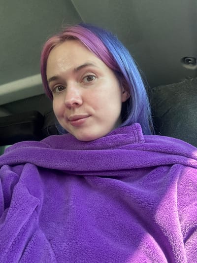 A woman is shown in a close-up, angled portrait. She has light skin, brown eyes, and her hair is dyed in two colors, with the roots near the top of her head colored blue, transitioning to a pink hue towards the tips. She is wrapped in a plush, purple robe. The background is indistinct but suggests an indoor setting. There is no information to suggest this photograph was taken in Le Muy, France.