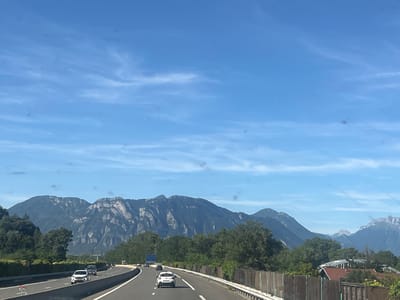 This is a highway scene in Barraux, France, on a clear day with a bright blue sky and scattered clouds. The perspective is from a moving vehicle, looking forward along a multi-lane highway. In the foreground, several cars are visible on the road, with a white car in the right lane being the most prominent. A wooden fence runs along the right side of the highway, and beyond that, residential buildings with red roofs are partially visible. The mid-ground features a dense line of green trees, and behind the trees, a majestic mountain range dominates the background. The mountains are rocky and rugged, with varying shades of green and gray. The sky is a vibrant blue, with thin, wispy clouds. The overall impression is of a scenic drive through the French countryside.