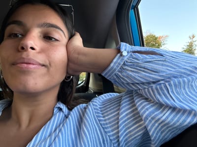 A single individual, a young adult female, is depicted seated inside a vehicle in the city of West Windsor, United States. She has light brown skin, dark brown hair pulled back, and dark eyes. She is wearing a light blue and white striped long-sleeved shirt, featuring a silver-colored circular button and a vertical slit on the cuff of her right sleeve. A silver-colored hoop earring is visible on her left ear. The frames of a pair of glasses are perched on top of her head. Her right hand is positioned with the palm against her right cheek and jawline, supporting her head. She is looking directly towards the camera with a neutral expression. The vehicle's interior is dark, with a blue door frame visible to the right of the subject. Outside the vehicle window, a clear blue sky and green trees are visible, indicating an outdoor setting during daylight hours.