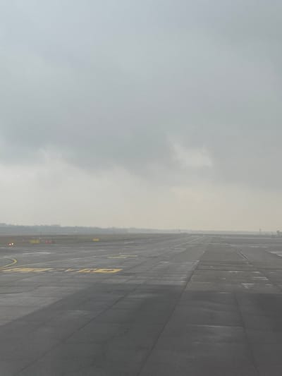 The scene depicts a section of an airport tarmac at Milan Malpensa Airport (MXP) in Somma Lombardo, Italy, under an overcast sky. The dark grey asphalt surface is wet, indicating recent precipitation, and features numerous yellow painted markings, including directional arrows, lines, and alphanumeric identifiers such as "A2" and "CM". In the mid-distance on the left, several orange-red airfield lights are illuminated along the edge of the tarmac. Further down the runway, a white aircraft, likely an airliner, is visible with its front lights illuminated. A hazy, dark green tree line stretches across the horizon, separating the airfield from the background. A faint, vertical structure, possibly a control tower or airport building, is minimally visible on the far right horizon. No individuals are present.