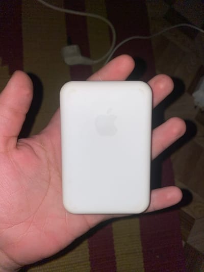 A white rectangular electronic device with an Apple logo is held in a person's hand. The device appears to be a portable charger or battery pack. The hand is positioned over a striped surface, likely a rug or fabric. In the background, a white charging cable and a white power adapter are visible, along with a dark, textured surface. The lighting is dim, suggesting an indoor environment. There is no indication of the city of Novosoldatka, Russia, or any forensic details that can be gleaned from the image.