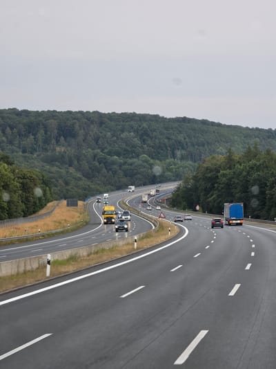 The image captures a view of a multi-lane highway in Bischbrunn, Germany, under an overcast sky. The highway curves through a landscape dominated by dense, green forests that rise on either side. Several vehicles are visible on the wet asphalt road, including a yellow truck in the foreground, a blue semi-trailer truck further ahead, and various cars and smaller trucks. The road is marked with white lines, indicating multiple lanes of traffic. Guardrails and roadside markers are present along the edges of the highway. The overall scene suggests a typical day of travel on a German autobahn, possibly during or after rainfall given the wet surface of the road and the diffused lighting conditions.