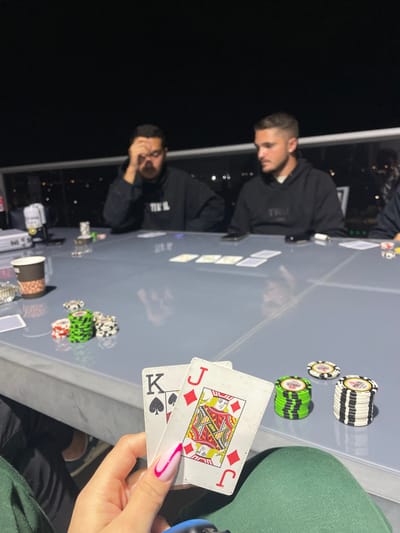 The image depicts a nighttime scene occurring on an elevated outdoor or semi-outdoor space in Yavne, Israel, with distant city lights visible against a dark sky. Three individuals are seated around a light grey rectangular table, participating in a card game.
In the foreground, a person's hand, featuring white manicured nails with a pink stripe, holds two playing cards: a King of Spades (K♠) and a Jack of Diamonds (J♦). This individual is wearing dark green trousers.
To the left of the table, a male subject with dark hair and a beard is seated, wearing a black hoodie. He is leaning forward with his right hand covering part of his face, his gaze directed downwards. To the center-right, another male subject with short dark hair is seated, also wearing a black hoodie; he is looking towards the left side of the table. A third subject is partially visible on the far right.
The table holds five face-up community cards in its center, indicating a poker game. Various stacks of poker chips (green, black, and white) are distributed on the table surface. Other objects include a brown paper cup, an ashtray, a white container, and a black smartphone in front of the center-right player. A light grey railing is visible in the background, behind the seated individuals. The scene is lit, highlighting the table and its occupants against the dark environment.