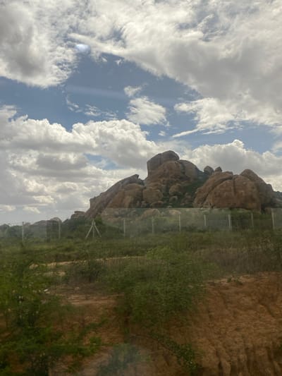 The image displays a vast natural landscape under a bright, partly cloudy sky, likely in Ihumwa, Tanzania. Dominating the right side of the scene are massive, weathered brownish-grey rock formations, resembling large boulders or kopjes.
A strip of green, bushy vegetation runs horizontally across the mid-ground, separating the rocky terrain from a drier, lighter-colored ground area. Within this vegetation, a series of slender, vertical structures, possibly fence posts or stakes, are partially visible, extending into the distance. The ground to the right of the vegetation appears to be dry, sandy earth.
The sky is a vivid blue, filled with numerous large, fluffy white cumulus clouds and some higher, more wispy cloud formations. This indicates clear, sunny daytime weather. There is no visible human activity or discernible subjects like people or animals in the frame. The scene is a static depiction of a natural environment, possibly taken from inside a vehicle due to a slight reflection across the image. No text is visible.