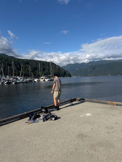 A young man stands on a concrete dock, facing away from the camera, overlooking a body of water. He is wearing a grey t-shirt and khaki shorts, with athletic shoes on his feet. In front of him on the dock are various fishing supplies, including a tackle box, a fishing rod, a blue water bottle, and a blue and black bag. Behind him, a marina is visible, filled with sailboats and other vessels. The water is calm and reflects the blue sky, which is dotted with white clouds. A dense forest of green trees covers the hills that rise behind the marina, and in the distance, mountain peaks are visible. The scene is set in Deep Cove, District of North Vancouver, Canada.