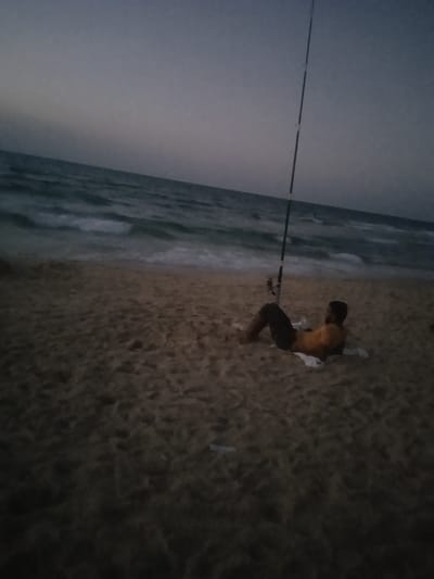 The image depicts a solitary figure on a sandy beach in Al-Mawasi, Palestinian Territories, during twilight or nighttime conditions. A single male individual is observed in the mid-ground, reclining on the sand, supported by his elbows with his knees bent. He is wearing a light-colored, short-sleeved shirt and dark pants, and is barefoot. A white cloth or towel is positioned beneath him. Directly in front of the individual, closer to the water's edge, a tall fishing rod is planted vertically in the sand, with its line extending into the ocean. The sea exhibits moderate waves, breaking onto the shore, with visible foam. The sky is dark grey, indicative of low light. Scattered light-colored debris is present on the sand in the foreground.