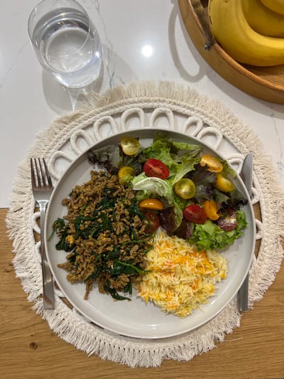 The image shows a plate of food on a white macrame placemat, placed on a table with a white countertop (likely marble) on one side and a wood surface on the other. The plate contains a salad with mixed greens and halved cherry tomatoes, rice with orange flecks, and ground meat mixed with spinach. A fork and knife are on either side of the plate. To the left, on the white countertop, is a glass of water and a wooden bowl filled with bananas. The setting suggests an indoor dining environment, possibly a home kitchen. The location in Groussbus-Wal, Luxembourg, is speculative, based solely on the possibility that this kind of meal setting and arrangement may be found in that geographical location.