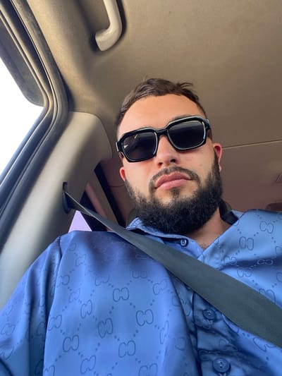 A man is seated in the passenger seat of a vehicle, looking at the camera. He is wearing black sunglasses and a blue button-down shirt with a repeating "GG" logo pattern. A black seatbelt is fastened across his chest. The interior of the car, including the headliner and a portion of the door frame, is visible in the background. The scene is lit by natural light coming from outside the vehicle. The location of Endirey, Russia is not depicted in this image.